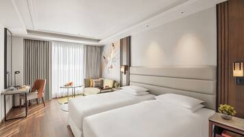 A modern hotel room with two beds, a desk, a chair, a sofa, and a large window with curtains.