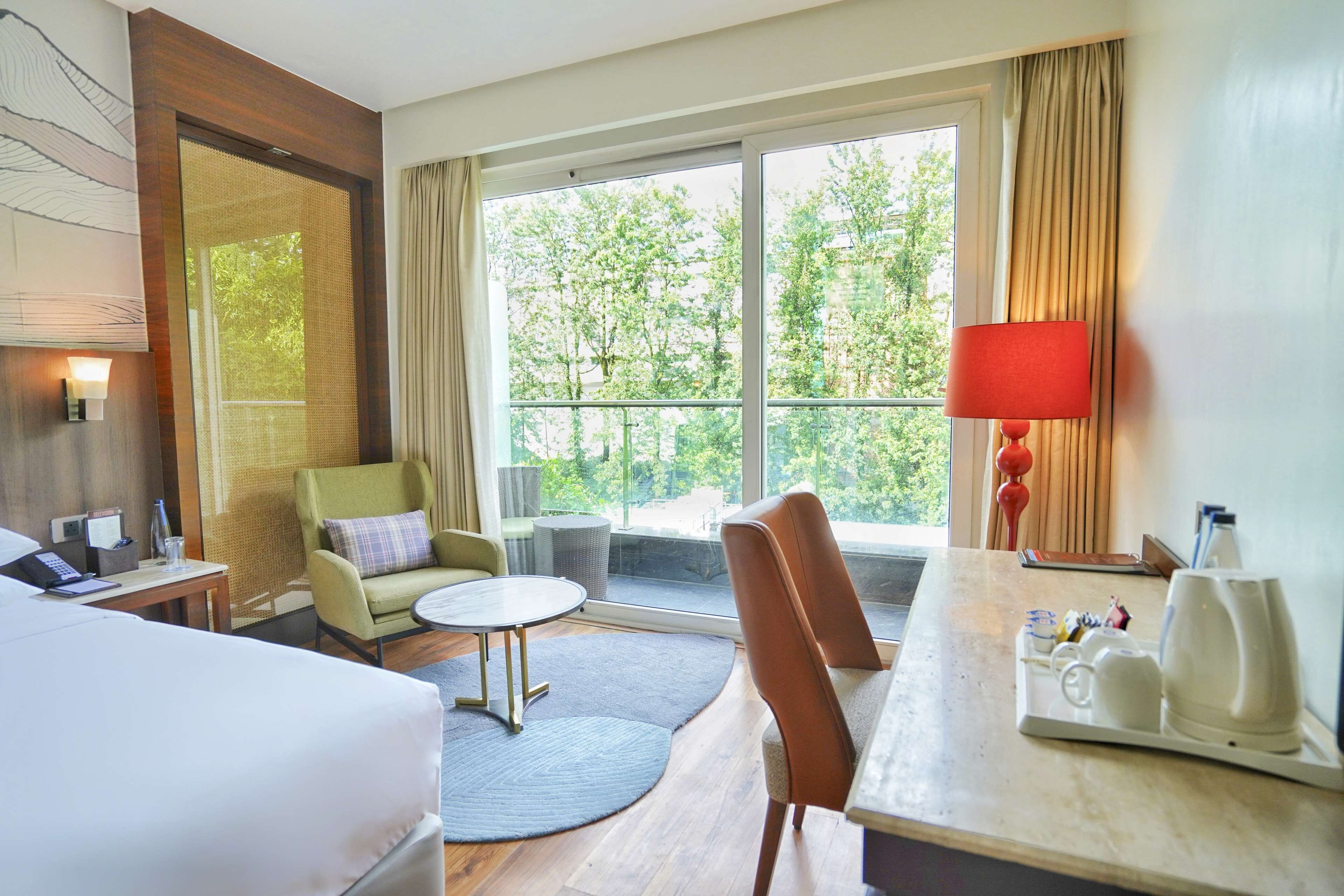 A hotel room with a bed, a desk with a lamp, a chair, a round table, and a view of trees through large windows.