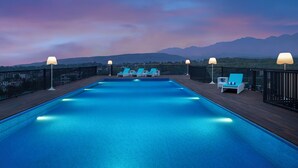 A swimming pool with lounge chairs and lamps, surrounded by a wooden deck and a metal fence, with mountains in the background. - Hyatt Centric Rajpur Road Dehradun (Dehradun)