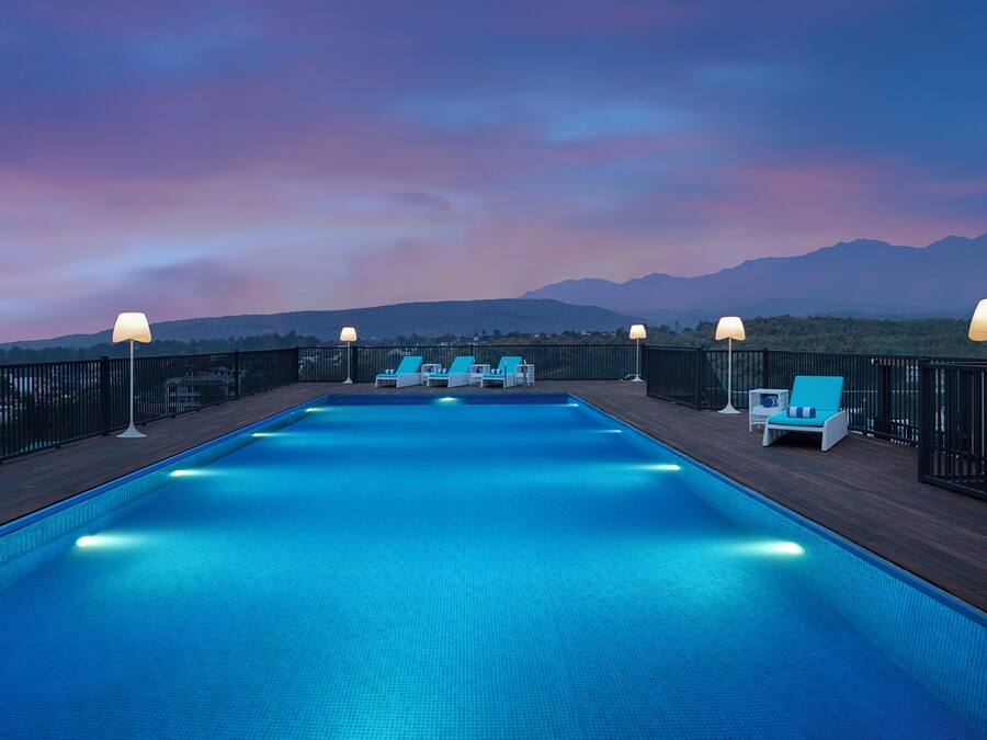 Una piscina con sillones de descanso y lámparas, rodeada por una terraza de madera y una valla de metal, con montañas al fondo.