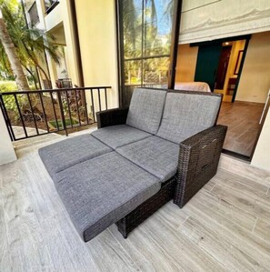 A grey sofa with a woven frame, positioned on a balcony with a view of a building and trees. - Pura Vida is activated (Guanacaste Province)