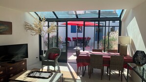 Una habitación iluminada por el sol con televisión, muebles de mimbre, una mesa de comedor con manteles rojos y vistas a un patio exterior con sombrillas rojas.