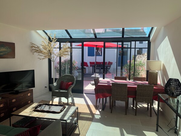 A sunlit room with a TV, wicker furniture, a dining table with red tablecloths, and a view of an outdoor patio with red umbrellas.