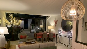 Una sala de estar moderna con comedor, que presenta una gran mesa de madera, un sofá verde y un juego de mesa con sillas. Hay un árbol decorativo con luces, una lámpara y un espejo montado en la pared.