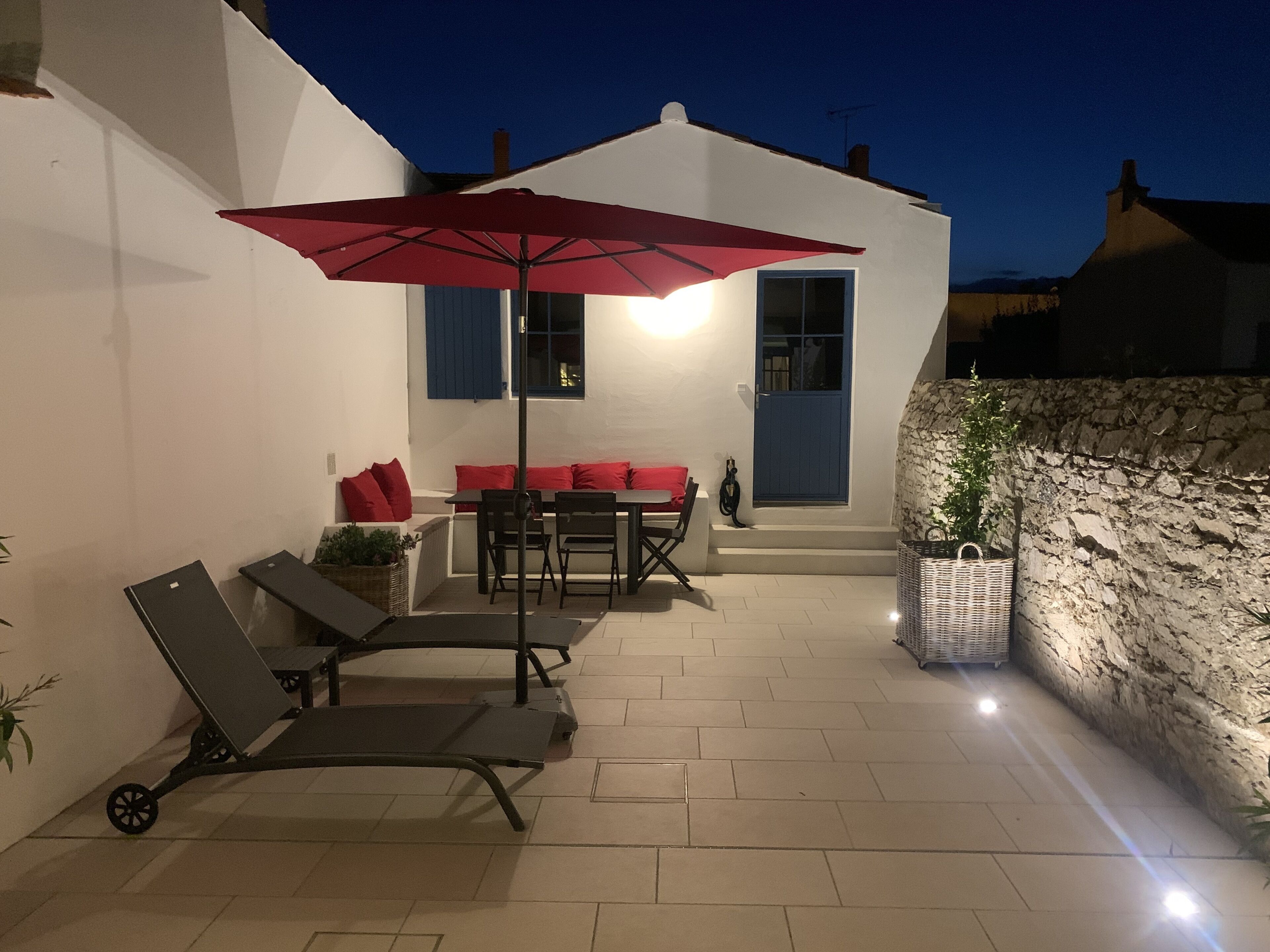 An outdoor patio with a table and chairs, a red umbrella, and a lounge chair.
