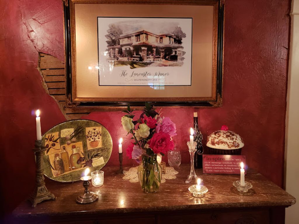 A framed picture of a house with a caption, a table with candles, a vase with flowers, and a sign with a quote.