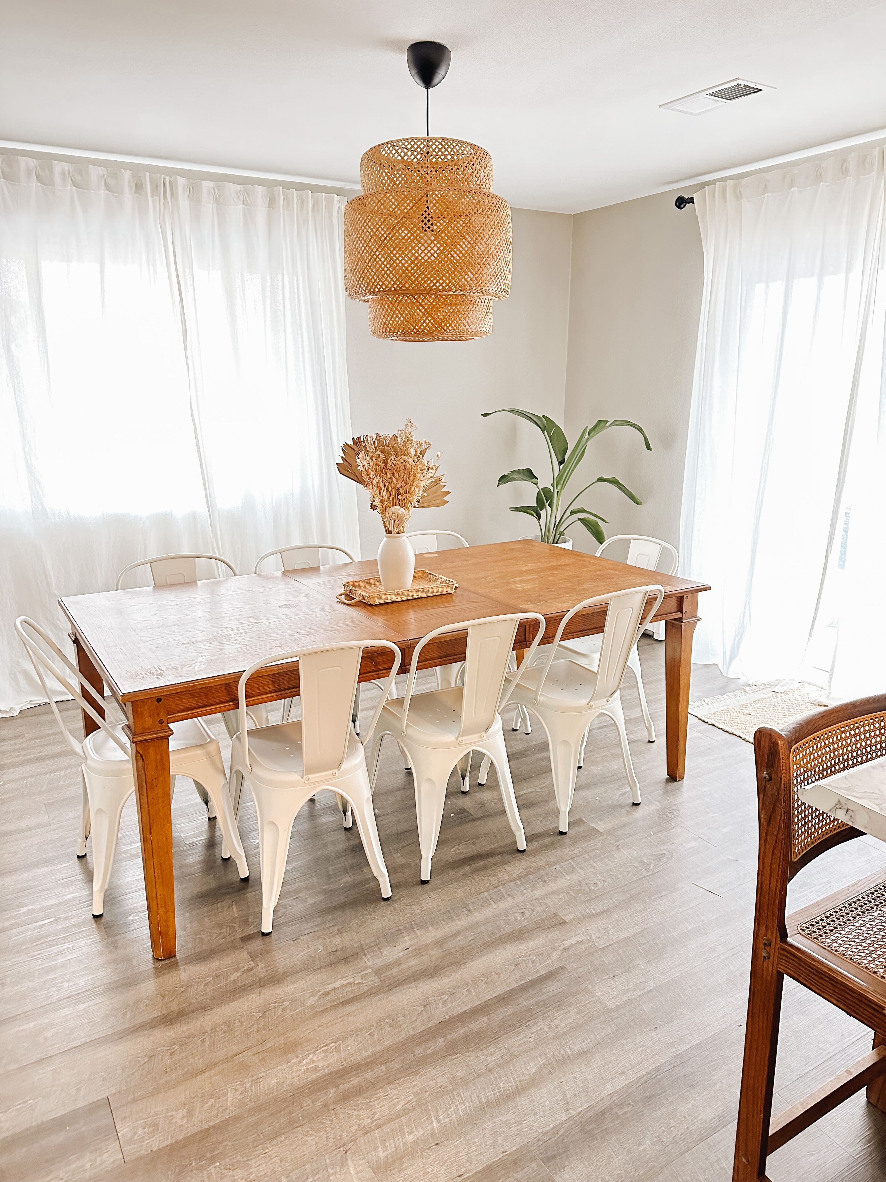 Un comedor con una mesa de madera y sillas blancas, una lámpara colgante de fibra y una planta en maceta.