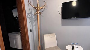 A room with a white chair, a small round table with a remote control, a coat rack, and a TV mounted on the wall. - Mazi Otel (Mudanya)