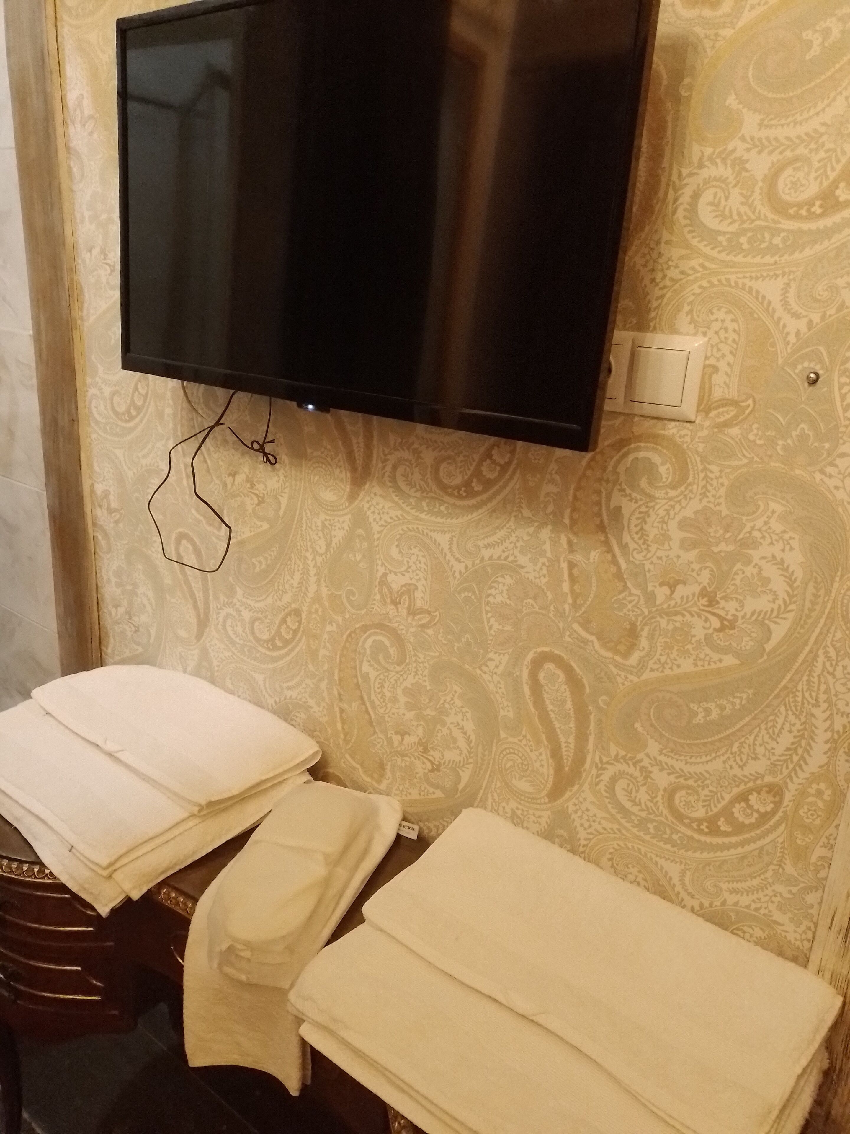 Una habitación con una televisión de pantalla plana en la pared, un papel tapiz estampado y una pila de toallas blancas sobre un banco de madera.