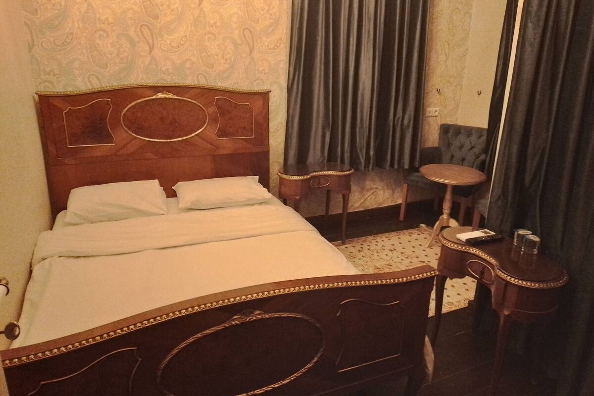 A hotel room with a wooden bed, a small round table, a chair, a mirror, and a patterned carpet.