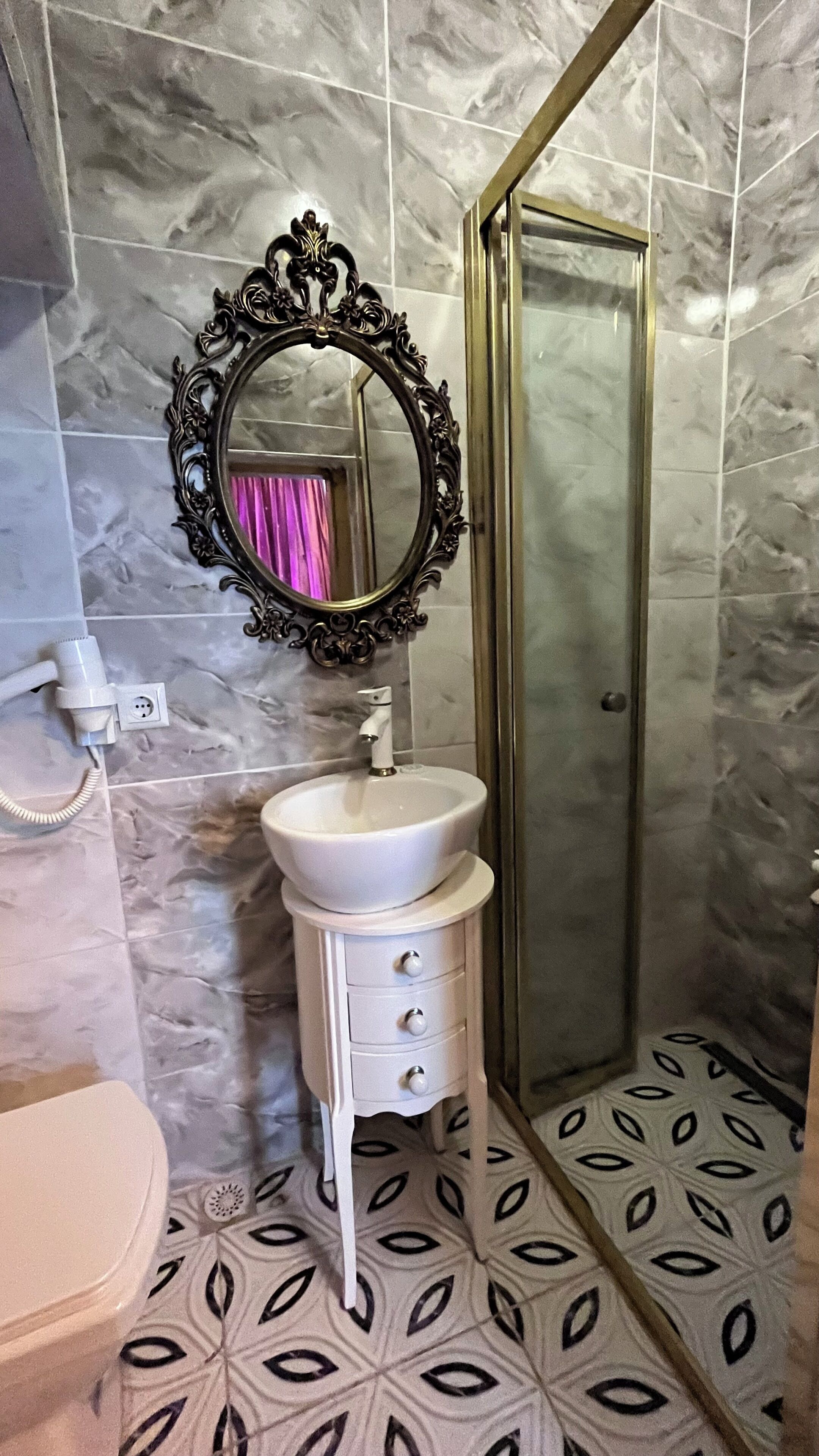 A bathroom with a white sink, a mirror with an ornate frame, a shower area, and patterned floor tiles.