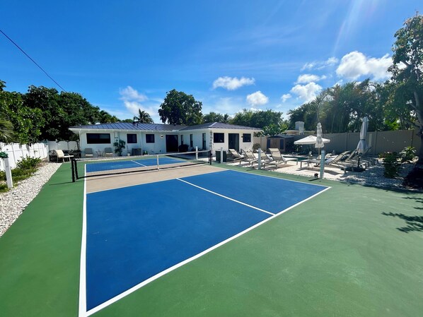 Una cancha de tenis con red, mobiliario exterior y un edificio al fondo.