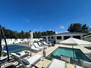Zona de piscina con sillas de descanso y sombrillas, cielo azul despejado y un edificio al fondo.