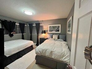Un dormitorio con dos camas, una mesita de noche con lámpara, un cuadro enmarcado en la pared y una ventana con persianas.