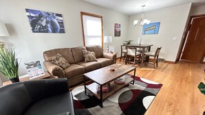 A living room with a brown leather sofa, a coffee table, a dining table with chairs, and a chandelier.