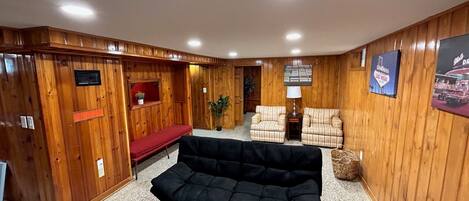 Una habitación con paneles de madera, un sofá negro, dos sillones estampados, un banco rojo y una mesa con un juego de mesa.