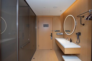 Un baño moderno con cabina de ducha de vidrio, tocador de madera con encimera blanca, espejo con luz y un perchero para toallas.