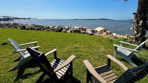Zona verde con varias sillas Adirondack orientadas hacia un cuerpo de agua, una orilla rocosa y un muelle a lo lejos.