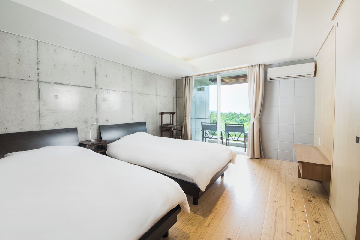 A modern hotel room with two beds, a concrete accent wall, a wooden floor, a balcony with a view, and a large window with curtains.