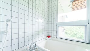A bathroom with white tiled walls, a bathtub, a showerhead, and a window with a view of greenery. - Asahi no Yado Shidakaji (Nanjo)