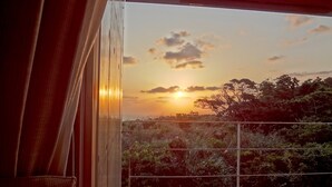 Ein Sonnenuntergang durch ein Fenster mit Vorhängen, einem Balkongeländer und dichtem Grün.