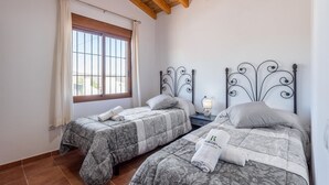 Una habitación con dos camas, cada una con cubrecama y almohadas estampadas, una mesita de noche con lámpara y una ventana con cortinas.