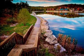A wooden boardwalk leading to a calm lake with a reflection of buildings and trees. - Book now for fall foliage! (Branson West)