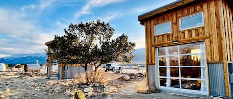 A rustic wooden cabin with large windows, surrounded by a rocky landscape and sparse vegetation.