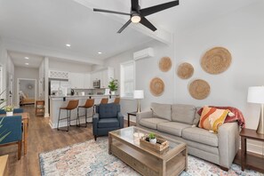 Una sala de estar moderna con sofá, mesa de centro y taburetes de bar. Se aprecia una zona de cocina con armarios blancos y un comedor con mesa y sillas.