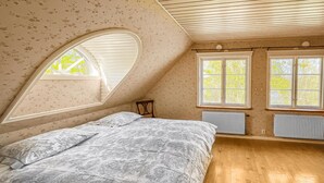 A bedroom with a sloped ceiling, a bed with patterned bedding, two windows with blinds, and a radiator.