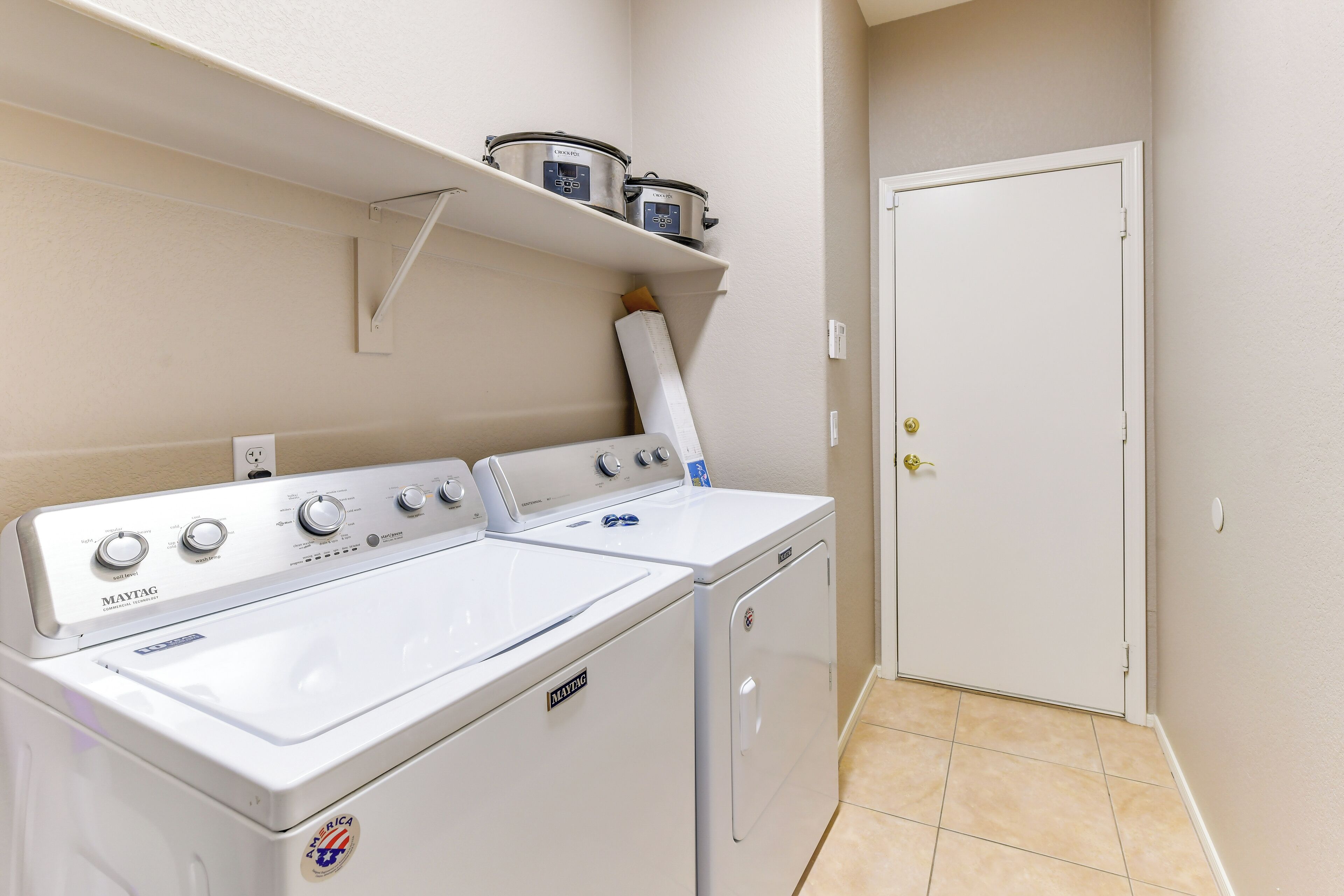 A laundry room with two Maytag washing machines, a shelf with a canister, and a white door.