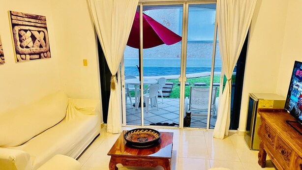 Sala de estar con un sofá blanco, una mesa de centro de madera y un televisor sobre un mueble de madera. Hay una puerta corrediza de vidrio que da a un área exterior con piscina y sillas.