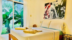 Un dormitorio con cama, una mesita de noche, una silla, un ventanal grande y un cuadro en la pared.