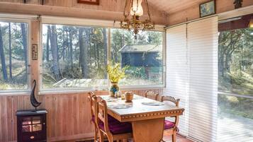 Une salle à manger en bois, avec une table dressée pour quatre personnes, un lustre et une vue sur une zone boisée grâce à de grandes fenêtres.