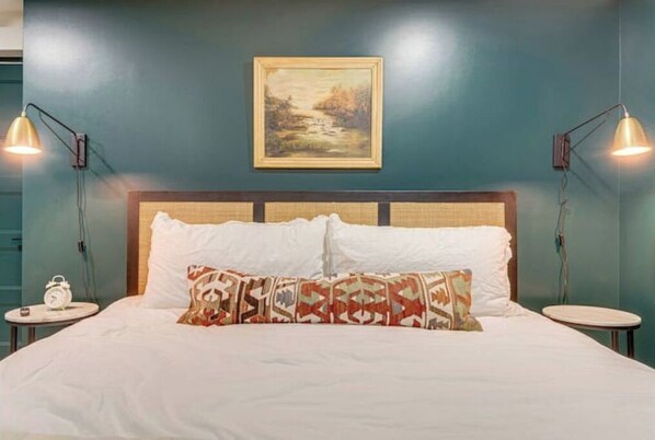 Un lit bien fait, agrémenté d’oreillers à motifs, une tête de lit ornée d’un motif tissé et un tableau représentant un paysage accroché au mur.