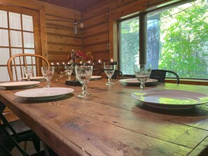 Une table à manger en bois, dressée avec des assiettes et de la verrerie, entourée de chaises, dans une pièce aux murs en bois et aux grandes fenêtres.