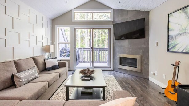 A spacious living room with a high ceiling, a fireplace, a large window, and a balcony. - Modern Ski-in Townhome with Hot Tub (Whistler)