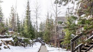 A snow-covered pathway flanked by trees and a building with a staircase. - Modern Ski-in Townhome with Hot Tub (Whistler)