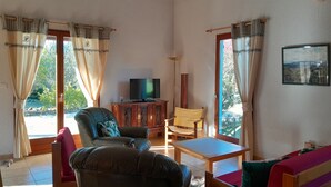 Un salon avec un canapé, un fauteuil, une table et un téléviseur. Il y a des rideaux aux fenêtres et un cadre avec une image accroché au mur.