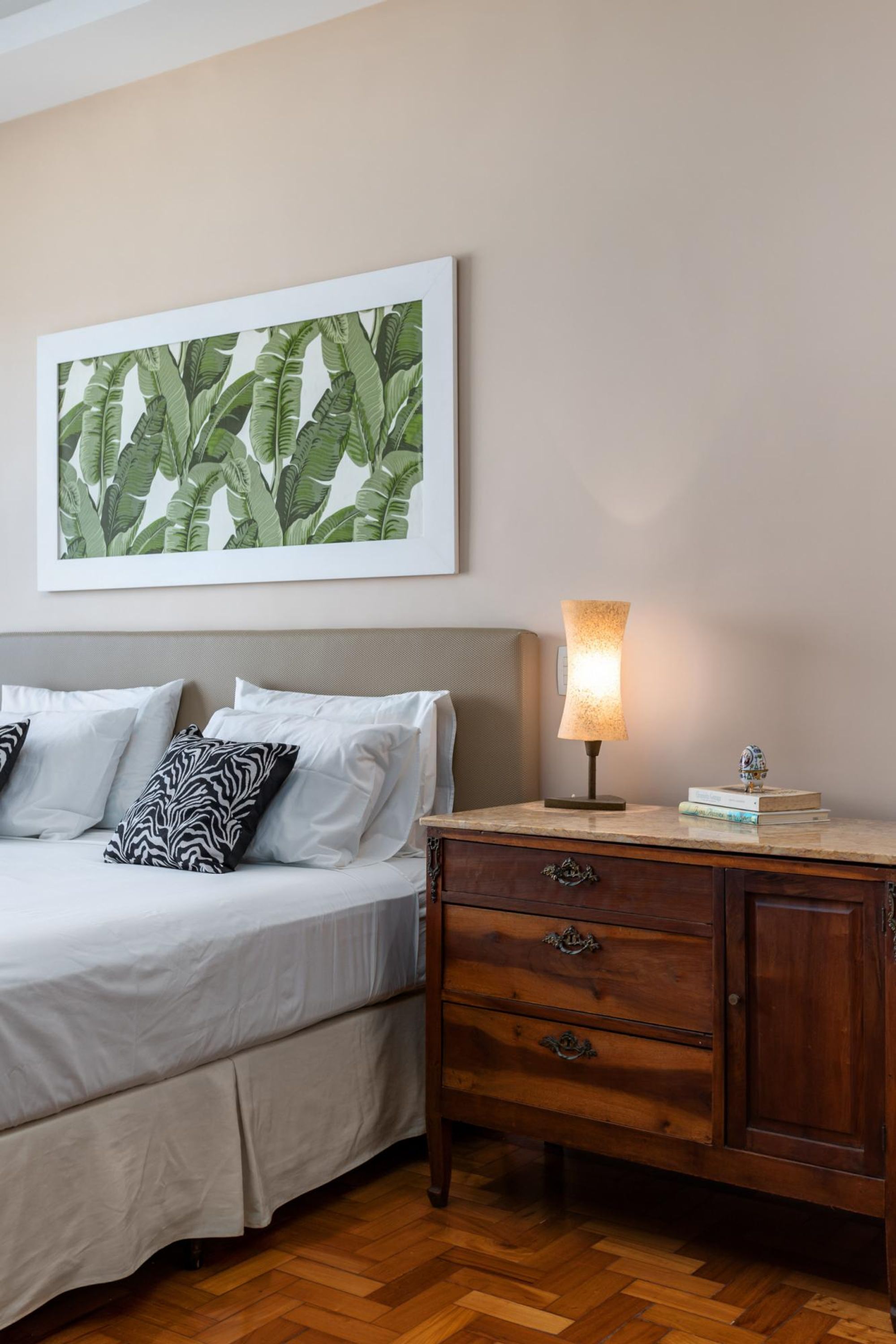 A bedroom with a bed, a nightstand with a lamp, a wooden dresser, and a framed botanical print on the wall.