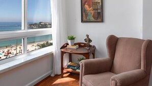 A room with a large window overlooking a beach, a brown armchair, a small wooden table with books and a plant, and a painting on the wall. - A 2 Minutos de la Playa de Copacabana A801 (Rio de Janeiro)