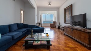 A living room with a blue sofa, a glass coffee table with books, a wooden cabinet, and a TV. - A 2 Minutos de la Playa de Copacabana A801 (Rio de Janeiro)