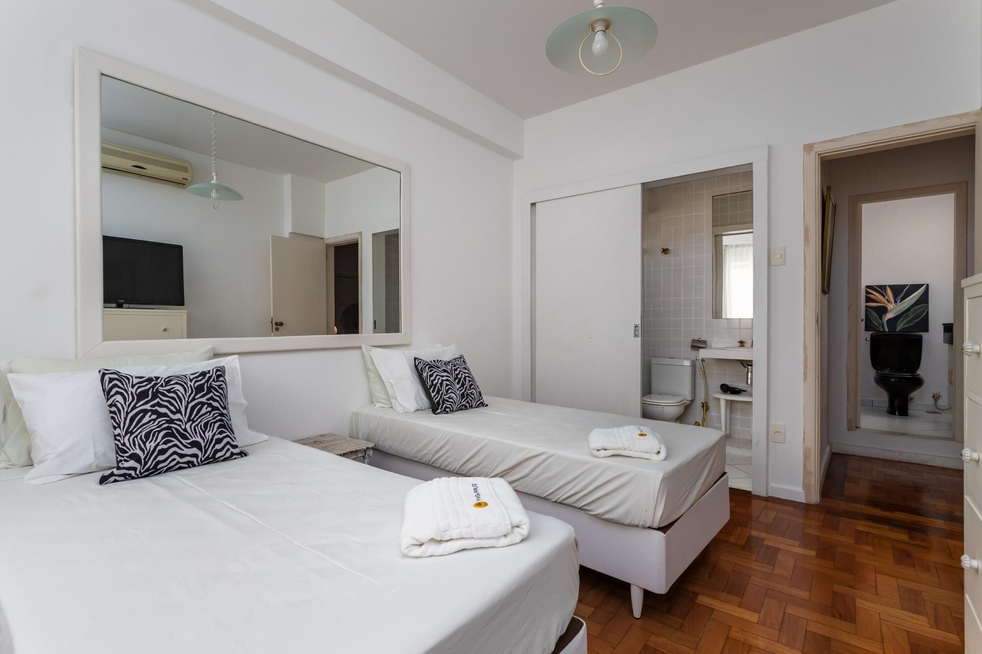 Camera d'albergo con due letti, un ampio specchio, una televisione e un bagno visibile attraverso una porta aperta.