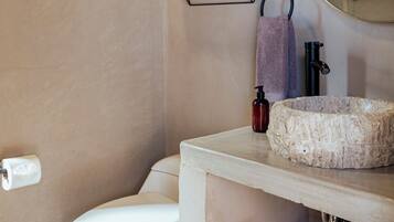 A bathroom with a white toilet, a wooden shelf with a towel and a bottle, a potted plant, and a round mirror.
