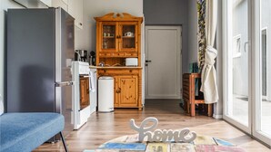 Eine kleine Küche mit Kühlschrank, Backofen und Holzschrank. Im Vordergrund sind eine Bank und ein kleiner Tisch zu sehen.