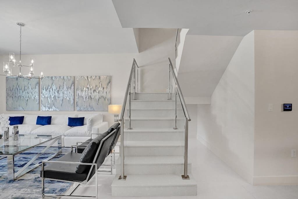 A modern living room with a glass coffee table, white sofa, and blue cushions. A staircase with a metal railing leads to an upper level.
