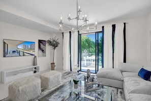 Un salon moderne avec une table basse en verre, des canapés blancs, un lustre et une grande fenêtre avec des rideaux.