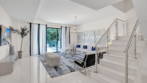 A modern living room with a glass coffee table, a sofa with blue cushions, a white ottoman, and a staircase with a glass railing. - Fort Lauderdale Luxury Condo (Fort Lauderdale)