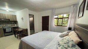 A bedroom with a bed, a nightstand, a window with curtains, a door, and a kitchenette with a stove and a chair. - Huanchacostudio/kingbed/balcony/tv/wifi (Huanchaco)