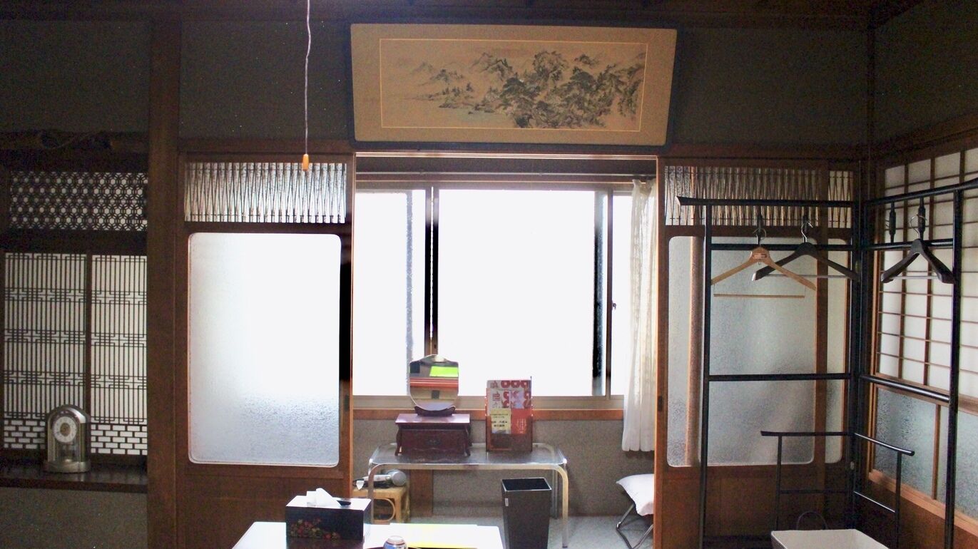 Een traditionele Japanse kamer met tatami vloerbedekking, een shoji schuifdeur, een futon bed, een klein tafeltje met een spiegel, een plank met boeken en een doos, en een kledingrek.